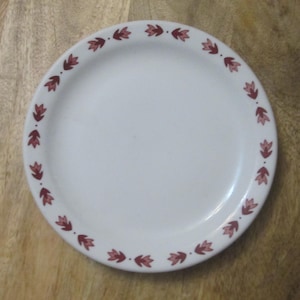 May include: A white ceramic plate with a decorative border of maroon floral designs. The plate is round and sits on a wooden surface. The floral pattern is evenly spaced around the rim.