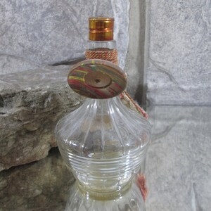 Puede incluir: Una botella de vidrio transparente con una superficie texturizada y un tap&oacute;n de madera. El tap&oacute;n est&aacute; pintado con rayas de colores y tiene un agujero en el centro. La botella tiene un borla que cuelga del tap&oacute;n.