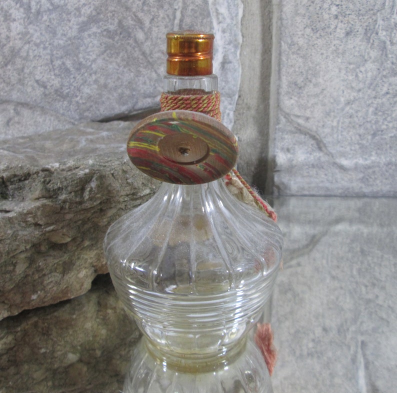 Puede incluir: Una botella de vidrio transparente con una superficie texturizada y un tap&oacute;n de madera. El tap&oacute;n est&aacute; pintado con rayas de colores y tiene un agujero en el centro. La botella tiene un borla que cuelga del tap&oacute;n.