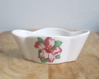 Op de afbeelding: Een kleine, crèmekleurige keramische kom met een geschulpte rand. De kom heeft een rood en roze bloemenontwerp met groene bladeren aan de voorkant. De kom staat op een houten oppervlak.