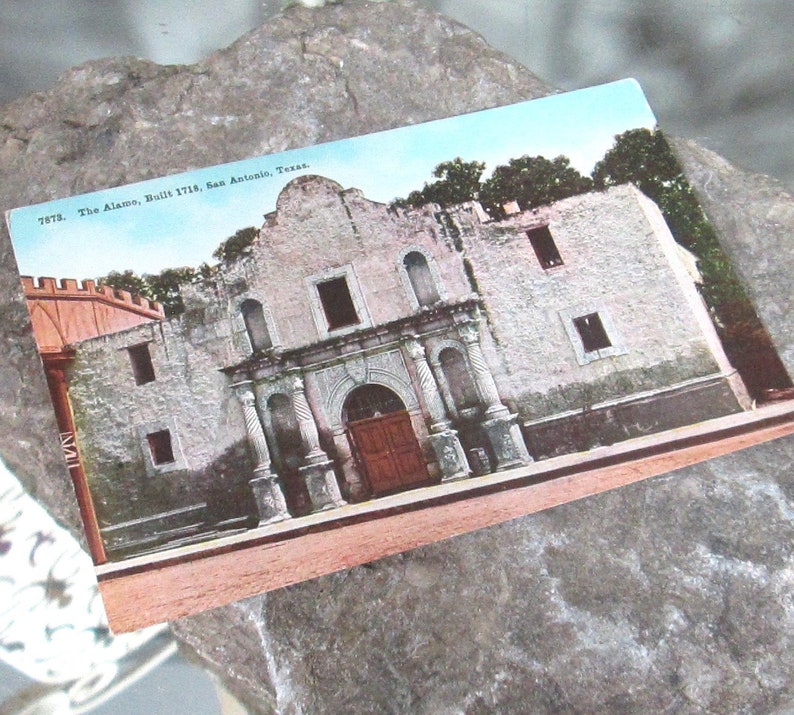 May include: A vintage postcard depicting The Alamo, a historic mission in San Antonio, Texas. The postcard features a photo of the building's facade, with its whitewashed walls, arched doorways, and small windows. The text on the postcard reads "7578. The Alamo. Built 1718. San Antonio, Texas."