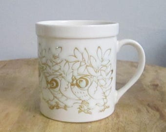 Könnte beinhalten: Weißer Keramikbecher mit Henkel, verziert mit einer goldenen Illustration von Eulen und Blättern. Der Becher hat eine zylindrische Form und einen breiten Rand. Das Eulendesign zeigt detaillierte Augen und Federn.