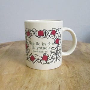 May include: Cream-colored ceramic mug with a handle. The mug features a design of red spools of thread and the text "Needle in the Haystack Pendleton, IN" in black. The design is framed by a decorative border.