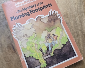 Könnte beinhalten: Ein Vintage-Buch mit dem Titel "The Mystery of the Flaming Footprints" von Alfred Hitchcock und The Three Investigators. Das Cover ist orange mit Illustrationen von Adlern und Figuren, mit dem Text "Terrifying events in a creepy old house!"