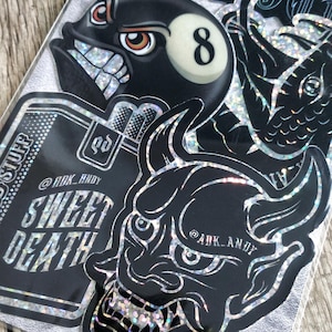 May include: A collection of holographic stickers featuring a black 8-ball character, a horned mask, and a rectangular design with the text SWEET DEATH and @ADK_ANDY. The stickers have a reflective, shimmering finish and bold black outlines.