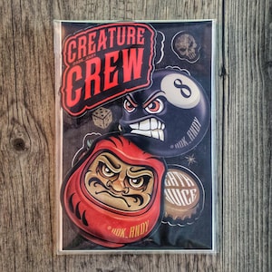 Könnte beinhalten: Ein rechteckiger Aufkleberbogen mit dem roten Schriftzug CREATURE CREW, einer schwarzen Acht-Ball-Kugel mit wütendem Gesicht, einer roten Daruma-Puppe, einem Würfel, einem Totenkopf-Symbol und einem braunen Kronkorken mit dem Text DEATH JUICE.