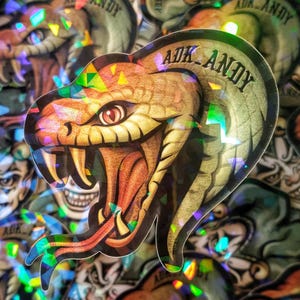 Op de afbeelding: Een holografische sticker met een gedetailleerde illustratie van een slang met open bek, die scherpe tanden laat zien. De sticker heeft een metallic glans en de tekst "ADK ANDY" is bovenaan gebogen. De achtergrond is gevuld met vergelijkbare stickers.
