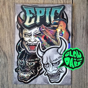 Könnte beinhalten: Eine Sammlung von Aufklebern mit dem Wort EPIC in Türkis und Schwarz sowie drei Hannya-Masken-Designs in Schwarz, Weiß und holografischen Oberflächen. Ein hellgrüner Sprechblasen-Aufkleber mit dem Text GLOW IN THE DARK ist enthalten.
