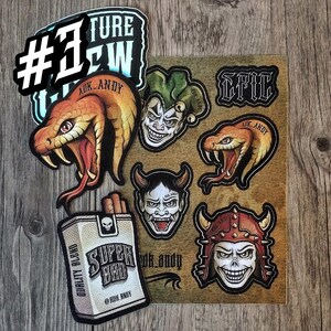 Può includere: Un foglio di adesivi con vari disegni grafici tra cui due serpenti arancioni, un giullare, un demone cornuto, un teschio con elmo chiodato e un pacchetto di sigarette con l'etichetta Super Bad. Il testo ADK_ANDY ed EPIC appare su diversi adesivi.