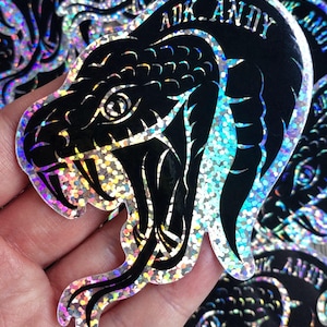 May include: A holographic sticker featuring a black snake head with an open mouth and visible fangs. The design includes the text ADK_ANDY arched above the snake. The background of the sticker has a reflective, multi-coloured glittery finish.