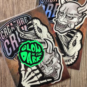 May include: A selection of stickers with diverse designs. One sticker has a green glow-in-the-dark design with the text "GLOW IN THE DARK". Other stickers include a snake, a demon, and a skeleton hand. The stickers have the text "CREATURE" and "CRI".