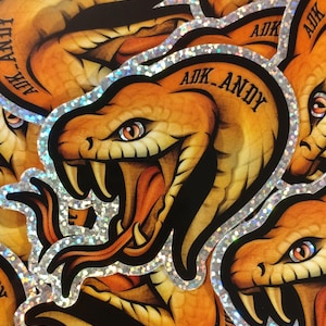 Op de afbeelding: Een verzameling stickers met een gedetailleerde illustratie van een slang met open bek, die tanden en een lange tong laat zien. De slang is voornamelijk oranje en bruin, met zwarte accenten en een zilveren glitterrand. De tekst "ADK_ANDY" is zichtbaar.