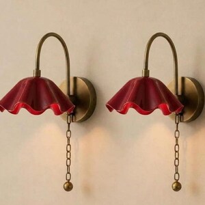 Puede incluir: Un par de lámparas de pared con brazos de latón curvados y placas circulares. Cada lámpara tiene una pantalla de color rojo intenso con forma de campana ondulada y una cadena de latón con un peso esférico al final.