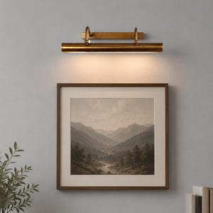 May include: A brass picture light fixture mounted above a square framed landscape painting. The painting depicts a valley with mountains, trees, and a river under a cloudy sky. The light fixture has a horizontal cylindrical shade and emits a warm glow.