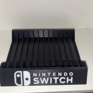 Può includere: Un supporto nero per cartucce di gioco Nintendo Switch. Il supporto ha più slot per cartucce di gioco e presenta il logo Nintendo Switch in bianco. Il supporto è realizzato in un materiale nero opaco.