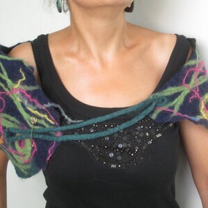 Könnte beinhalten: Ein blauer, grüner und rosa gefilzter Wollschal mit einem gelben Blumenakzent. Der Schal ist um die Schultern gebunden und mit einer Sicherheitsnadel befestigt.