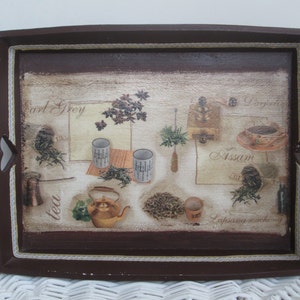 May include: A rectangular brown serving tray with a decorative tea-themed design. The tray features images of tea leaves, teacups, a teapot, and text labels like "Earl Grey" and "Assam". The tray has cut-out handles and a white lace trim.
