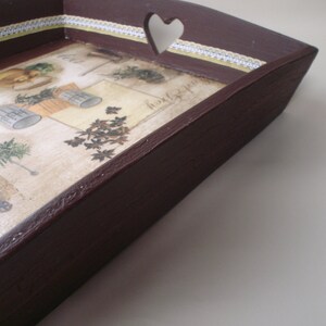 May include: A dark brown wooden serving tray with a heart-shaped cutout on one side. The tray's interior features a vintage-style botanical print with herbs and gardening tools. A decorative white and yellow lace trim borders the print.