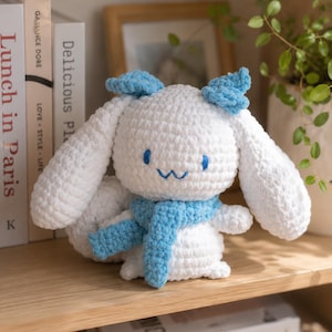 Peut inclure: Une peluche lapin blanche au crochet avec des accents bleus, notamment des oreilles en forme de nœud et une écharpe. Le lapin a des yeux bleus et un sourire cousu. Il est exposé sur une étagère en bois avec des livres et une plante en arrière-plan.