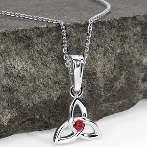Può includere: Una collana con pendente color argento caratterizzata da un design a nodo triquetra con una piccola pietra rossa rotonda e sfaccettata incastonata al centro. Il pendente pende da una delicata catenina argentata, con una finitura metallica lucida.