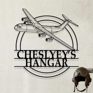 May include: Black metal airplane wall art with the text "CHESLYEY'S HANGAR" in a bold font. The airplane is a detailed outline, set within a circular frame. A vintage aviator helmet is visible on the right.