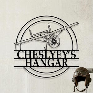 以下が含まれることがあります： 円形のフレームにプロペラ機のシルエットと「CHESLYEY'S HANGAR」の文字が描かれた黒い金属製の壁掛け看板。看板の横には、ヴィンテージデザインのゴーグル付き茶色のレザー製飛行帽があり、顎紐が付いています。