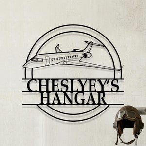 May include: Black metal wall art featuring a detailed outline of a private jet inside a circular frame. The text "CHESLYEY'S HANGAR" is displayed below the jet. A vintage aviator helmet is visible on the right.