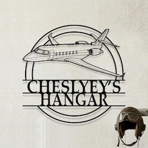 Puede incluir: Arte mural de metal negro con un contorno detallado de un jet privado dentro de un marco circular. El texto "CHESLYEY'S HANGAR" se muestra debajo. Un casco de aviador vintage con gafas está a la derecha.