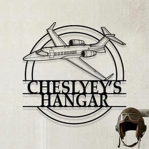 May include: A black metal wall sign featuring a silhouette of a private jet flying over a circular frame. The text "CHESLYEY'S HANGAR" is displayed in bold, capitalized letters across the center of the design, integrated into the circular structure.