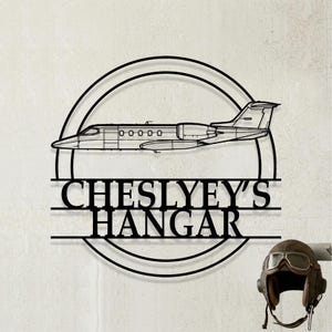 May include: A black metal wall sign featuring a circular frame with a silhouette of a private jet airplane in the center. Below the airplane, the text CHESLYEY'S HANGAR is displayed in bold, capital letters, integrated into the circular design.