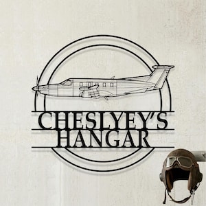 Könnte beinhalten: Ein schwarzes Metall-Wandschild mit einem kreisförmigen Rahmen und der Silhouette eines Flugzeugs im Seitenprofil in der Mitte. Unter dem Flugzeug steht der Text CHESLYEY'S HANGAR in fetten Großbuchstaben. Ein brauner Fliegerhelm aus Leder liegt daneben.