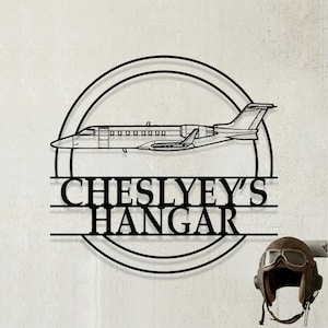 May include: A black metal wall sign featuring a circular frame with a silhouette of a private jet airplane in the center. Below the airplane, the text reads CHESLYEY'S HANGAR in bold capital letters. A brown leather aviator cap is positioned nearby.