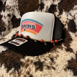 May include: A black and white trucker hat with the San Antonio Spurs logo on the front. The hat features a white front panel with the team name and logo in red, orange, and blue. A decorative chain with basketball and star charms adorns the brim.