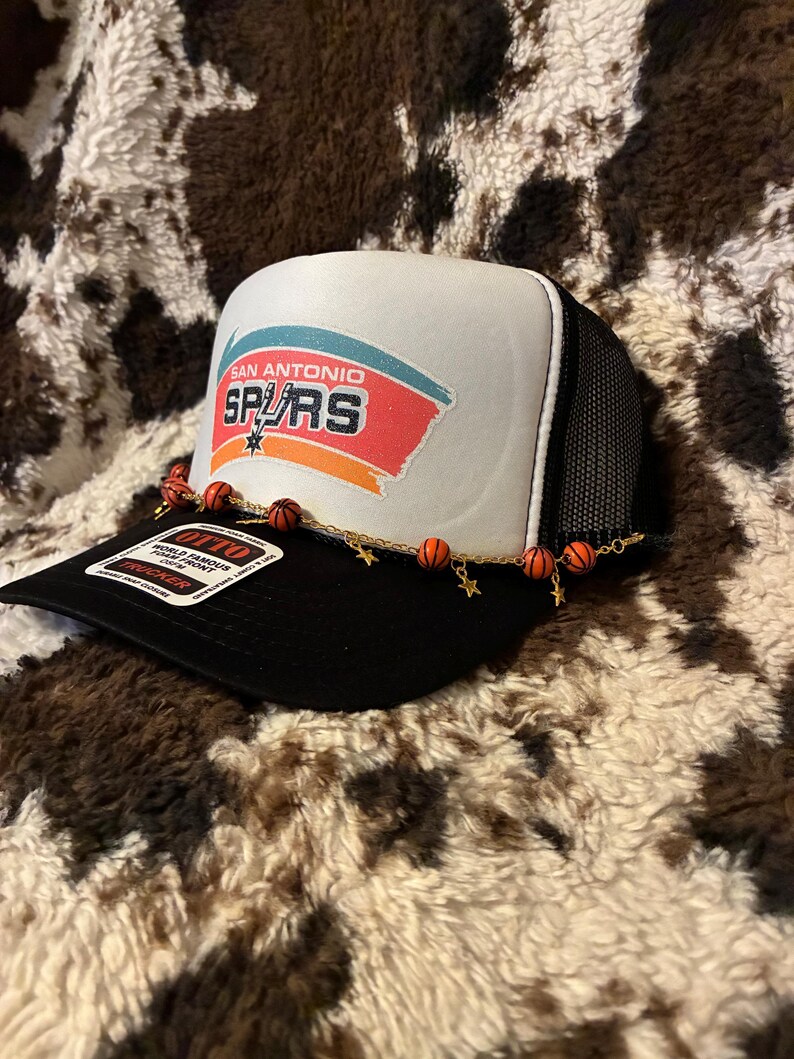 May include: A black and white trucker hat with the San Antonio Spurs logo on the front. The hat features a white front panel with the team name and logo in red, orange, and blue. A decorative chain with basketball and star charms adorns the brim.