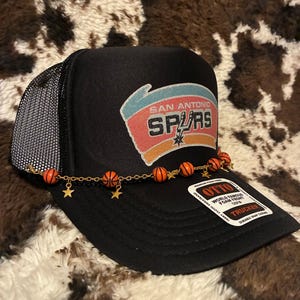 May include: Black trucker hat featuring the San Antonio Spurs logo in red, orange, and white. The hat has a mesh back and a decorative chain with basketball and star charms. The hat has a curved brim and a snap closure.