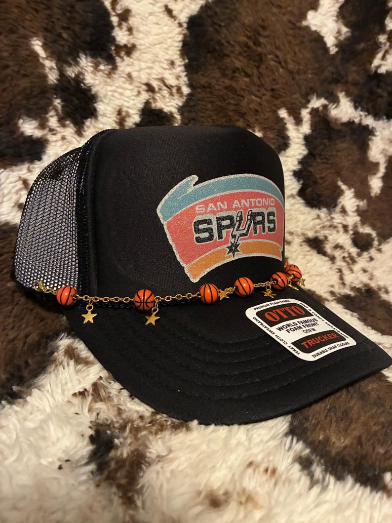May include: Black trucker hat featuring the San Antonio Spurs logo in red, orange, and white. The hat has a mesh back and a decorative chain with basketball and star charms. The hat has a curved brim and a snap closure.