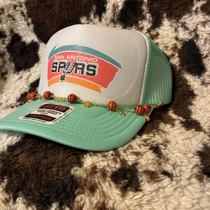 May include: A San Antonio Spurs trucker hat with a white front panel and a mint green mesh back. The hat features the team logo and a decorative chain with basketball beads and gold star charms. The hat is displayed on a brown and white patterned surface.