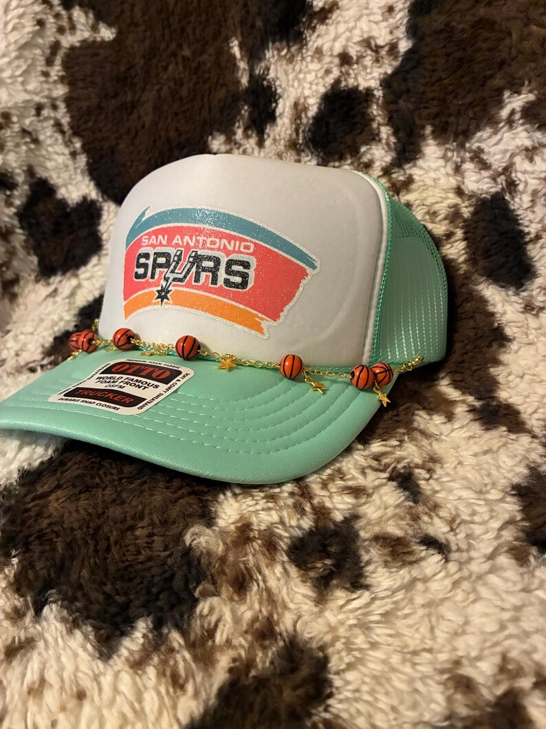 May include: A San Antonio Spurs trucker hat with a white front panel and a mint green mesh back. The hat features the team logo and a decorative chain with basketball beads and gold star charms. The hat is displayed on a brown and white patterned surface.