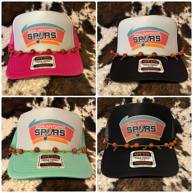 May include: Four San Antonio Spurs trucker hats in pink, black, mint green, and black. Each hat features the team logo on the front, a mesh back, and a decorative chain with basketball charms. The hats have the text "SAN ANTONIO SPURS" and "OTTO TRUCKER" on the front.