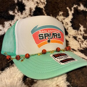 May include: A white and mint green trucker hat with the San Antonio Spurs logo. The hat features a mesh back, a curved brim, and a decorative chain with basketball and star charms. The hat has the text "OTTO" and "TRUCKER".