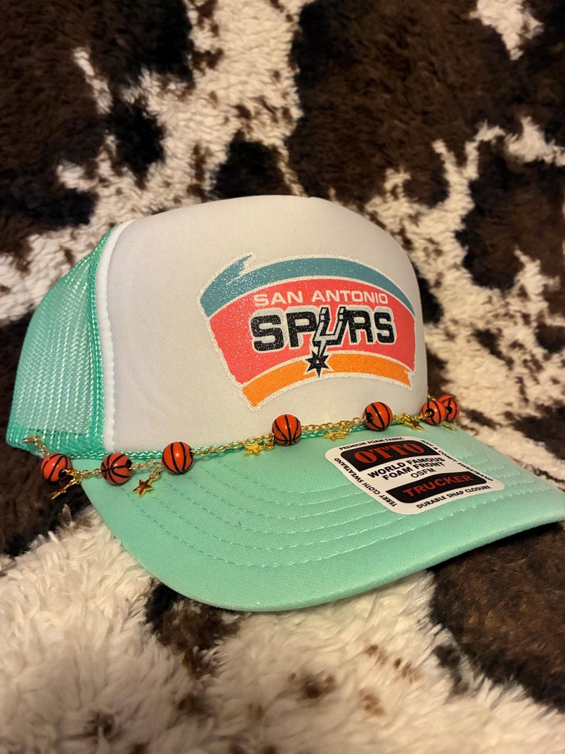 May include: A white and mint green trucker hat with the San Antonio Spurs logo. The hat features a mesh back, a curved brim, and a decorative chain with basketball and star charms. The hat has the text "OTTO" and "TRUCKER".