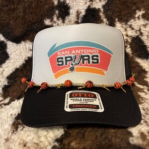 May include: Black and white trucker hat with the San Antonio Spurs logo in the center. The logo features the team name and a star. The hat has a black brim and a white front panel. A decorative chain with small orange basketballs is attached to the brim.