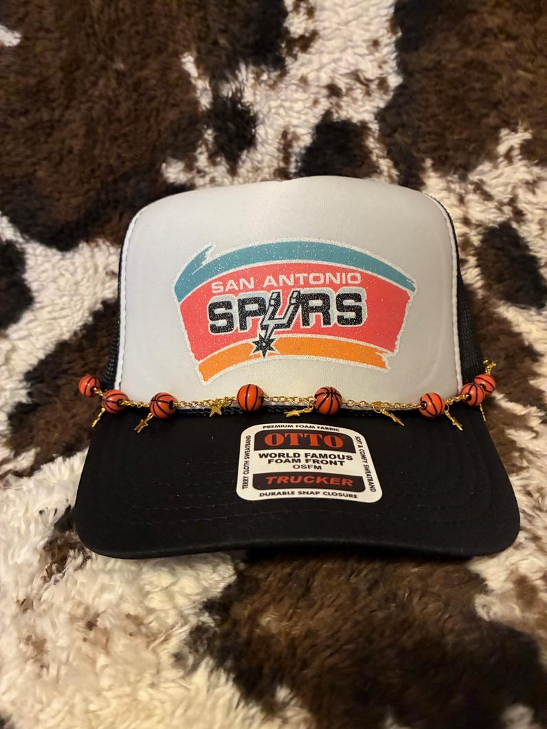 May include: Black and white trucker hat with the San Antonio Spurs logo in the center. The logo features the team name and a star. The hat has a black brim and a white front panel. A decorative chain with small orange basketballs is attached to the brim.