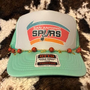 May include: A mint green and white trucker hat featuring the San Antonio Spurs logo. The hat has a mesh back and a curved brim. A decorative chain with basketball beads adorns the front. The hat has the text "OTTO" and "TRUCKER".