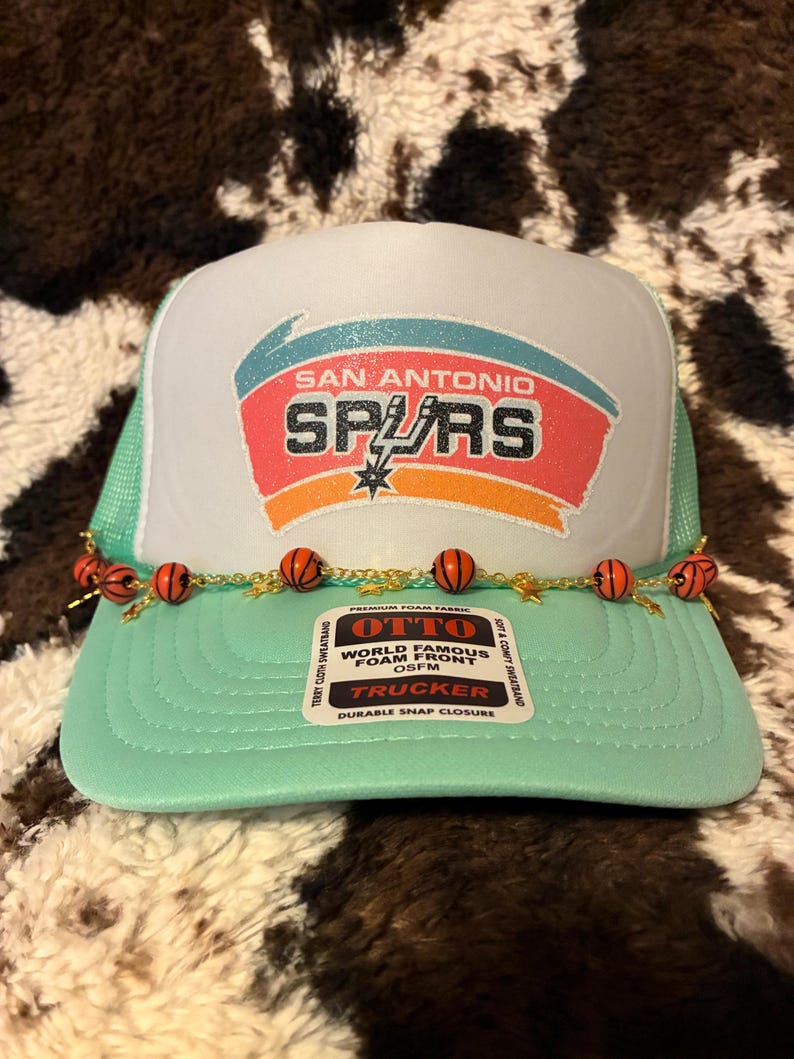 May include: A mint green and white trucker hat featuring the San Antonio Spurs logo. The hat has a mesh back and a curved brim. A decorative chain with basketball beads adorns the front. The hat has the text "OTTO" and "TRUCKER".