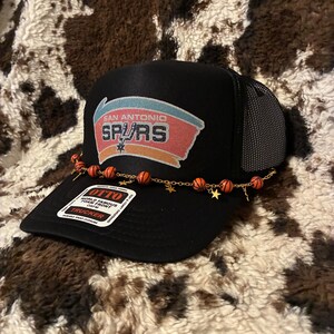 May include: Black trucker hat with the San Antonio Spurs logo on the front. The hat has a mesh back and a curved brim. A gold chain with basketball beads and star charms is attached to the brim. The tag reads "OTTO WORLD FAMOUS FOAM FRONT TRUCKER".