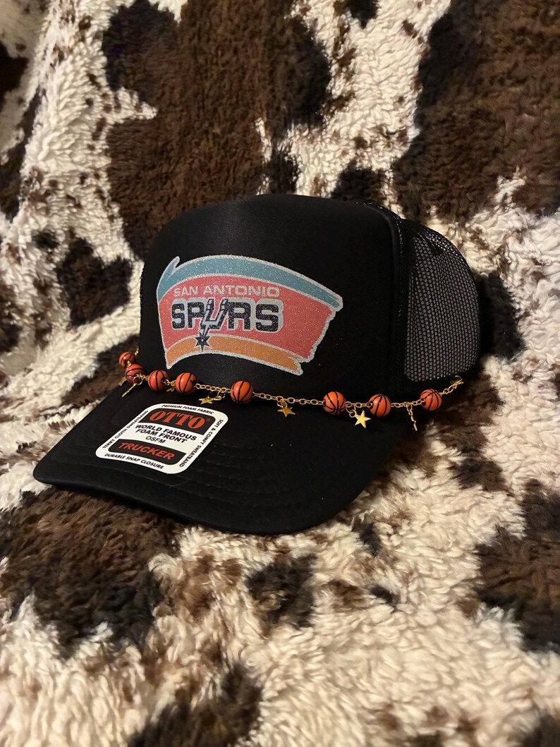 May include: Black trucker hat with the San Antonio Spurs logo on the front. The hat has a mesh back and a curved brim. A gold chain with basketball beads and star charms is attached to the brim. The tag reads "OTTO WORLD FAMOUS FOAM FRONT TRUCKER".