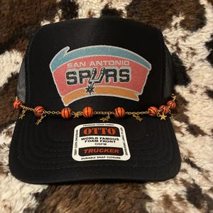 May include: Black trucker hat featuring the San Antonio Spurs logo in red, orange, and white. The hat has a mesh back and a decorative chain with basketball and star charms. The hat is labeled "OTTO" and "TRUCKER".