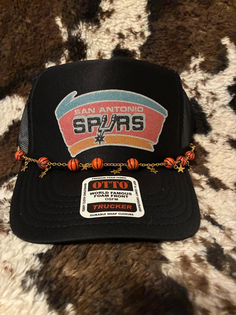 May include: Black trucker hat featuring the San Antonio Spurs logo in red, orange, and white. The hat has a mesh back and a decorative chain with basketball and star charms. The hat is labeled "OTTO" and "TRUCKER".