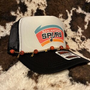 May include: A black and white trucker hat with the San Antonio Spurs logo on the front. The hat features a mesh back, a curved brim, and a decorative chain with basketball and star charms.